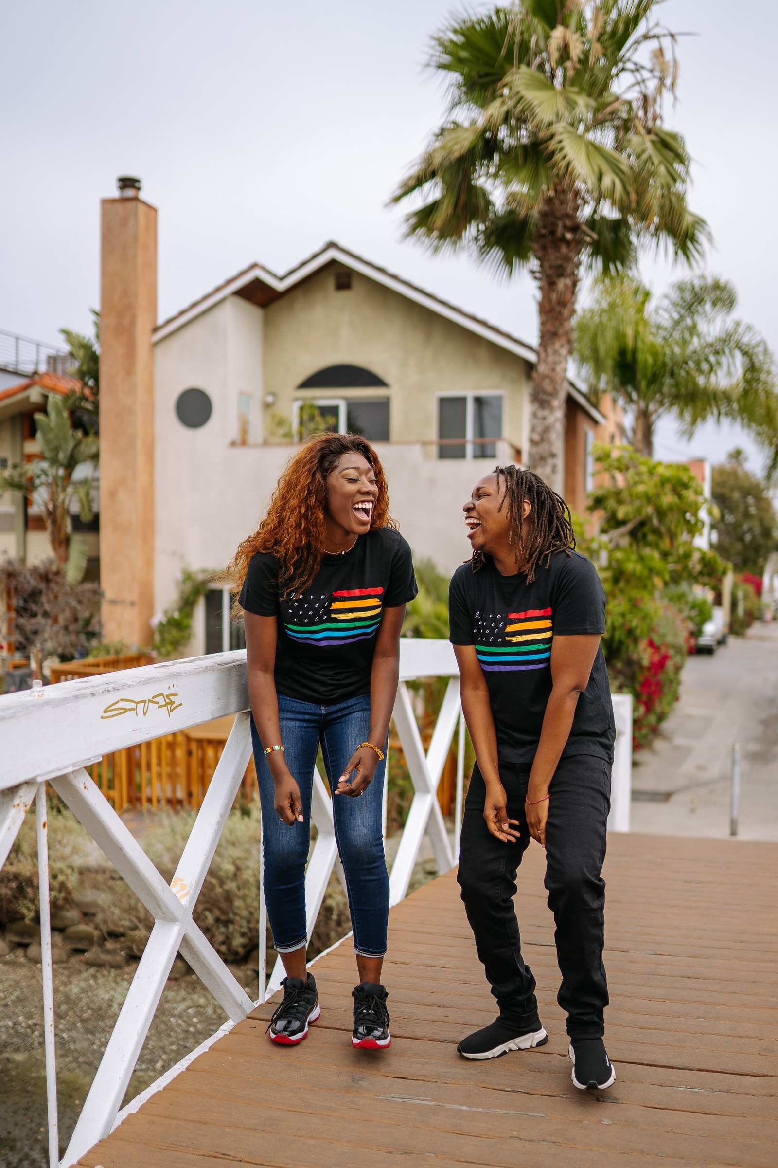 Two young Black friends laughing together on a wooden footbridge, both wearing black T-shirts printed with rainbow pride stripes.