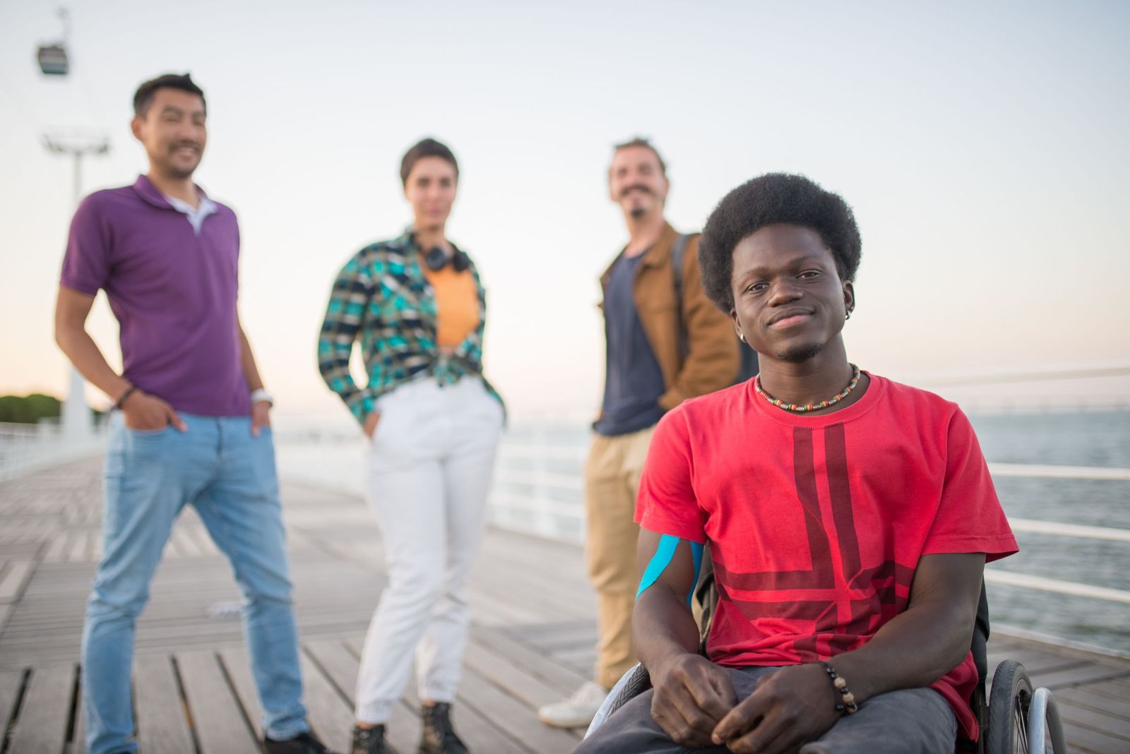 A young Black man using a wheelchair smiles at the camera on a waterfront boardwalk at dusk, with three friends standing out of focus behind him.