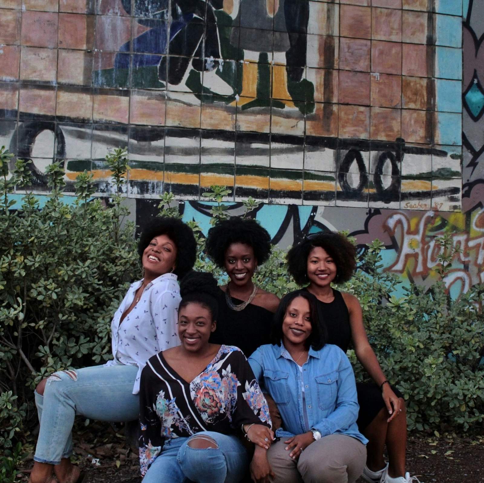 Five Black women smiling together in front of a colourful mural